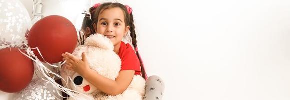 linda niño pequeño niña posando con rojo corazón conformado globos y un osito de peluche oso aislado en blanco. niños Moda foto