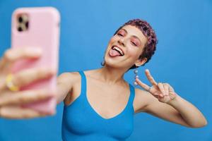 Young sports woman fashion blogger takes a picture of herself on the phone in blue sportswear smiling and showing her tongue on a blue monochrome background photo