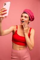 A young woman blogger with colored pink hair and a short haircut takes a picture of herself on the phone and broadcasts a smile in stylish clothes and a hat on a pink background monochrome style photo