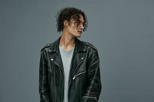 Handsome stylish tanned curly man leather jacket looks aside posing isolated on over gray studio background. Cool fashion offer. Huge Seasonal Sale New Collection concept. Copy space for ad photo