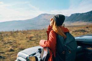 woman in a jacket with a backpack and a cap stands near the car on nature in the mountains in summer photo