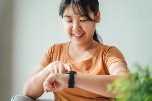 joven mujer mirando y utilizando un reloj inteligente, hembra sentado en el sofá comprobación su reloj inteligente. foto