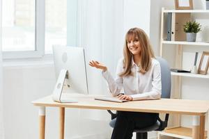 Smiling cheerful adorable blonde businesswoman worker freelancer look at camera raise hand up in light modern office. Happy employee work on computer online, enjoy successful business. Copy space photo