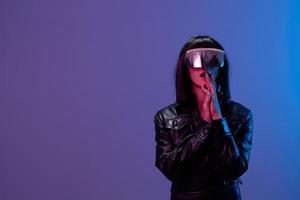 Scared shocked awesome brunet woman in leather jacket trendy specular sunglasses fold hands near face posing isolated in blue violet color light background. Neon party Cyberpunk concept. Copy space photo