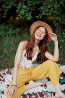 Hippie woman smiling in eco clothing yellow pants, white knit top, hat and yellow glasses sitting on plaid in park watching sunset, lifestyle camping trip photo