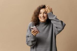 Overjoyed lucky curly cute lady in gray casual sweater get cool news from message posing isolated on over beige pastel background. Social media, network, distance communication concept. Copy space photo