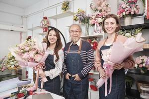 Portrait of flower shop workers team. Asian old male florist owner and young beautiful female employees in aprons with bunch of fresh floral bouquets arrangement for SME business, happy work in store. photo