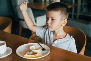 pequeño y linda chico teniendo desayuno en cafetería. comiendo panqueques con agrio crema foto