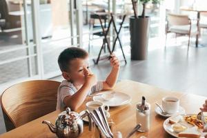 pequeño y linda chico teniendo desayuno en cafetería. comiendo panqueques con agrio crema foto