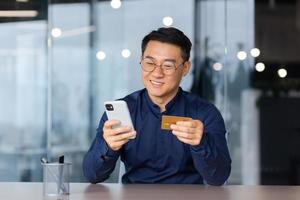 Satisfied and smiling man inside office at work, using app on phone and bank credit card for online shopping in online store, money transfer and booking, close up asian man with smartphone photo