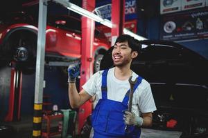 retrato de sonriente asiático masculino reparador participación con las piernas cruzadas llave inglesa a garaje profesional reparar y mantenimiento. coche inspección Servicio foto