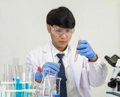 asiático masculino estudiante científico vistiendo un del doctor vestido en el laboratorio mirando mano a químico. causado por mezcla reactivos en científico investigación laboratorios con prueba tubos y microscopio en el mesa foto