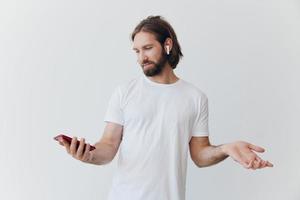 A man with a hipster beard in a white T-shirt with a phone and wireless headphones smiling listening to music and an audiobook online against a white wall photo