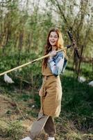 un mujer granjero en trabajo ropa y un delantal trabajos al aire libre en naturaleza y sostiene un rastrillo a reunir césped foto