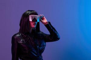 Excited awesome brunet woman in leather jacket touch trendy specular sunglasses open mouth look aside posing isolated in blue violet color light background. Neon party Cyberpunk concept. Copy space photo