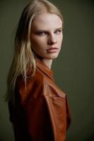 Closeup studio portrait of blonde with expressive glance blue eyes showy makeup looking over her shoulder at camera. Beautiful young woman in trendy leather brown dress posing isolated on green wall photo