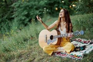 joven hembra hippie artista obras de teatro guitarra y canta canciones en Respetuoso del medio ambiente ropa sentado en el suelo fuera de en naturaleza en el otoño mirando fuera a el puesta de sol foto