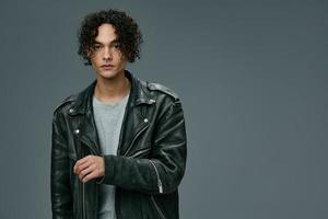 Handsome fashionable stylish tanned curly man leather jacket posing isolated on over gray studio background. Cool fashion offer. Huge Seasonal Sale New Collection concept. Copy space for ad photo