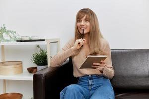 Enjoyed blonde student lady with copybook smiles at camera sitting in sofa couch at modern living room interior. Female client have shrink appointment in a psychological help office. Copy space photo