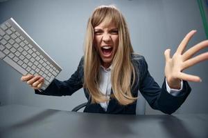 Crazy irritated angry millennial blonde businesswoman worker in blue jacket throws keyboard and screaming at camera in gray modern office. People Emotions Business Concept. Copy space, wide angle photo