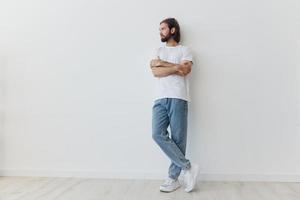 A man with a beard and long hair in a white T-shirt and blue jeans stands against a white wall leaning full-length on it, relaxed style photo