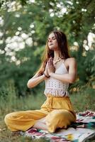 A young hippie woman meditates in nature in the park, sitting in a lotus position on her colorful plaid and enjoying harmony with the world in eco-clothing photo