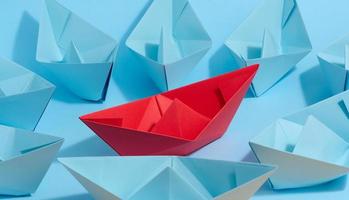 A red paper boat stands in front of a group of blue paper boats, a confrontation photo