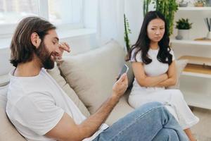 A man looks at the phone screen during an argument with his girlfriend. The angry and hurt woman looks in his direction and is sad. Family discord at home, phone addiction photo