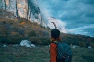 un viajero con un mochila y en un rojo suéter muestra el montañas con su mano y el azul cielo nubes foto