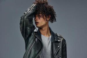 Adorable stylish tanned curly man leather jacket touches hair looks aside posing isolated on gray studio background. Cool fashion offer. Huge Seasonal Sale New Collection concept. Copy space for ad photo