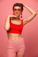 Young athletic woman with a short haircut and purple hair in a red top and pink yoga leggings in sunglasses with an athletic figure smiles and dances on a pink background photo