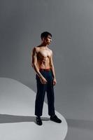 a handsome man with a naked torso in jeans and shoes stands on a gray background in full growth photo