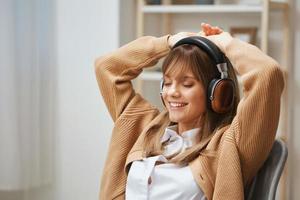 Happy young blonde lady freelancer in warm sweater in headphones listen moves to favorite songs dancing sitting in armchair at home interior. Music time Relaxing Cool playlist Concept. Copy space photo