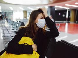 pasajero vistiendo un médico máscara sentado a el aeropuerto amarillo mochila esperando foto