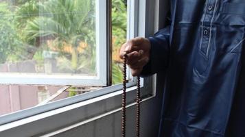 A young man was standing holding tasbih in typical Muslim clothing by the window. photo