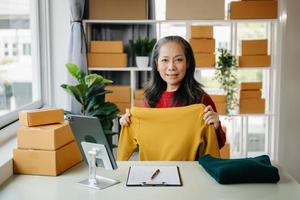 Fashion blogger concept, Senior Asian women selling clothes on video streaming.Startup small business SME, using smartphone or tablet taking receive and checking photo