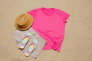 Sand beach texture background. Mockup pink summer t-shirt copy space. Blank template woman shirt Top view. Summertime accessories hat, flip-flops. Flat-lay closeup tshirt on seashore. Beachtime photo