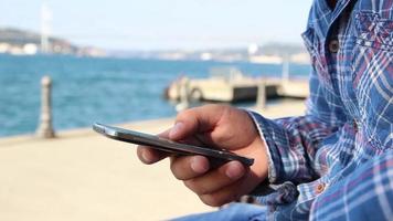 Person verwenden Smartphone und Sitzung mit ein Sicht, Meer Sicht, Brücke, im ein sonnig Tag, selektiv Fokus, Lärm bewirken video
