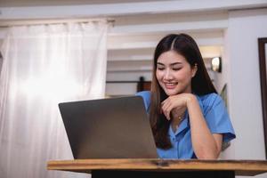 Asian woman with a beautiful smile and face sits comfortably on her desk at home with a laptop and notebook for relaxing and relaxing in a bright morning. holiday vacation concept. photo