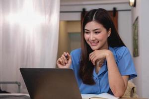 Asian woman with a beautiful smile and face sits comfortably on her desk at home with a laptop and notebook for relaxing and relaxing in a bright morning. holiday vacation concept. photo
