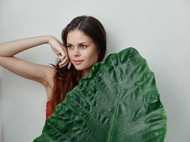 red-haired woman holds hand near face green leaf Exotic swimsuit Studio photo