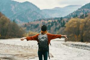 woman in a red sweater cap with a backpack on the river bank and mountains in the distance photo