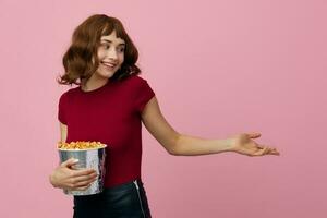 Smiling cheerful pretty cute redhead lady in red shirt with popcorn ready to watch movie posing isolated on over pink studio background. Copy space Banner. Fashion Cinema concept. Entertainment offer photo