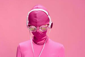 Art portrait of a woman wearing a pink full-face burglar mask with glowing round glasses wearing pink clothes with pink headphones on a pink background looking into the camera photo