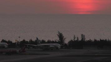 PHUKET, THAILAND FEBRUARY 11, 2023 - Tractor pulls low cost airline AirAsia on the airfield of Phuket airport. Airplane silhouette at sunset background. Airport by the sea. Tourism and travel concept video