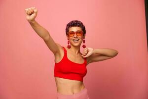 un joven atlético mujer con un corto Corte de pelo y púrpura pelo en un rojo parte superior en Gafas de sol con un atlético figura sonrisas con su brazos extendido en diferente direcciones bailando en un rosado antecedentes foto