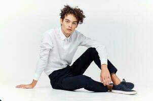 curly-haired guy in shirt and trousers and sneakers sits on the floor indoors side view photo