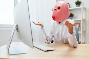 Angry funny crazy businesswoman worker freelancer in pink fish mask work on computer online pondering or making decision shocked by operational problems spread hands in light modern office. Copy space photo