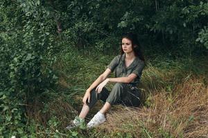 woman in overalls and sneakers sits on the grass in the forest photo