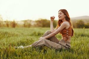 A woman sits on the green young fresh grass in the open air and laughs and smiles happily in the sunset light, her flying red hair fluttering in the wind photo